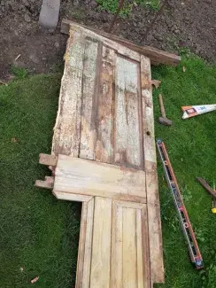 an old wooden panelled door laying on the ground for repair with two upper panels and one lower right panel intact but the lower left panel missing along with the left side. there are some remnants of the old paint